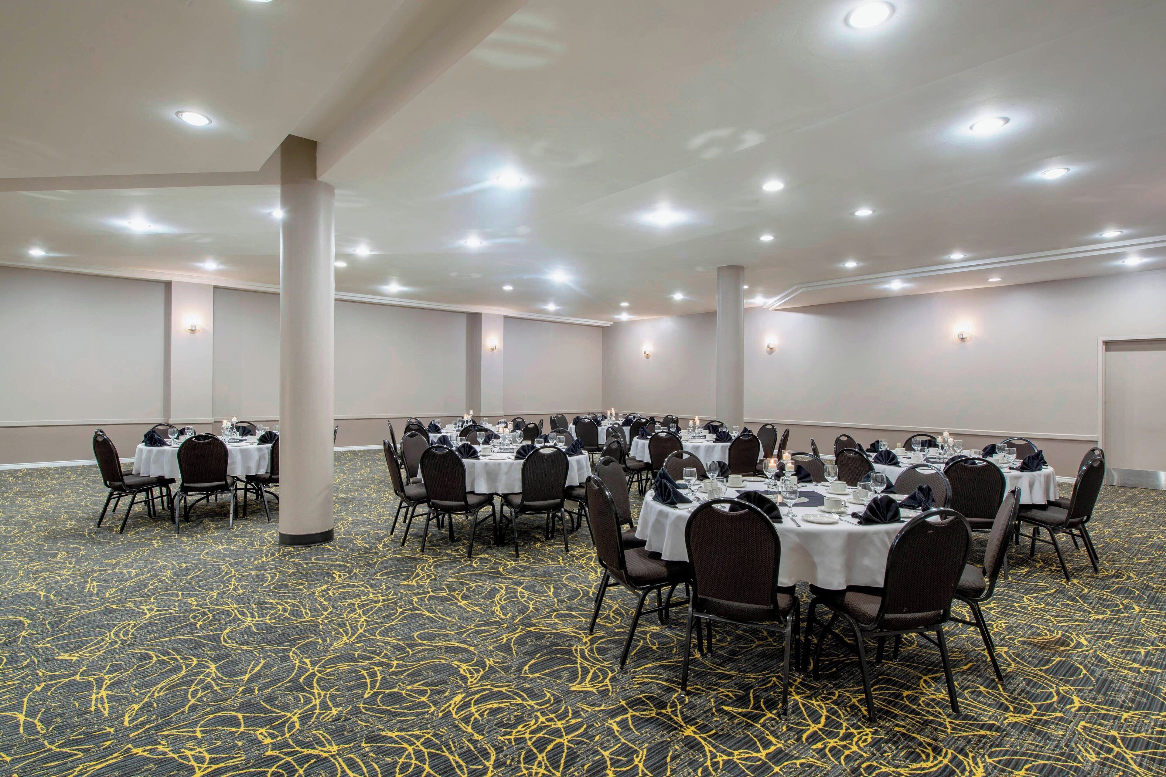 A conference room with multiple tables and chairs, arranged in rows, with a carpeted floor and ceiling lights.