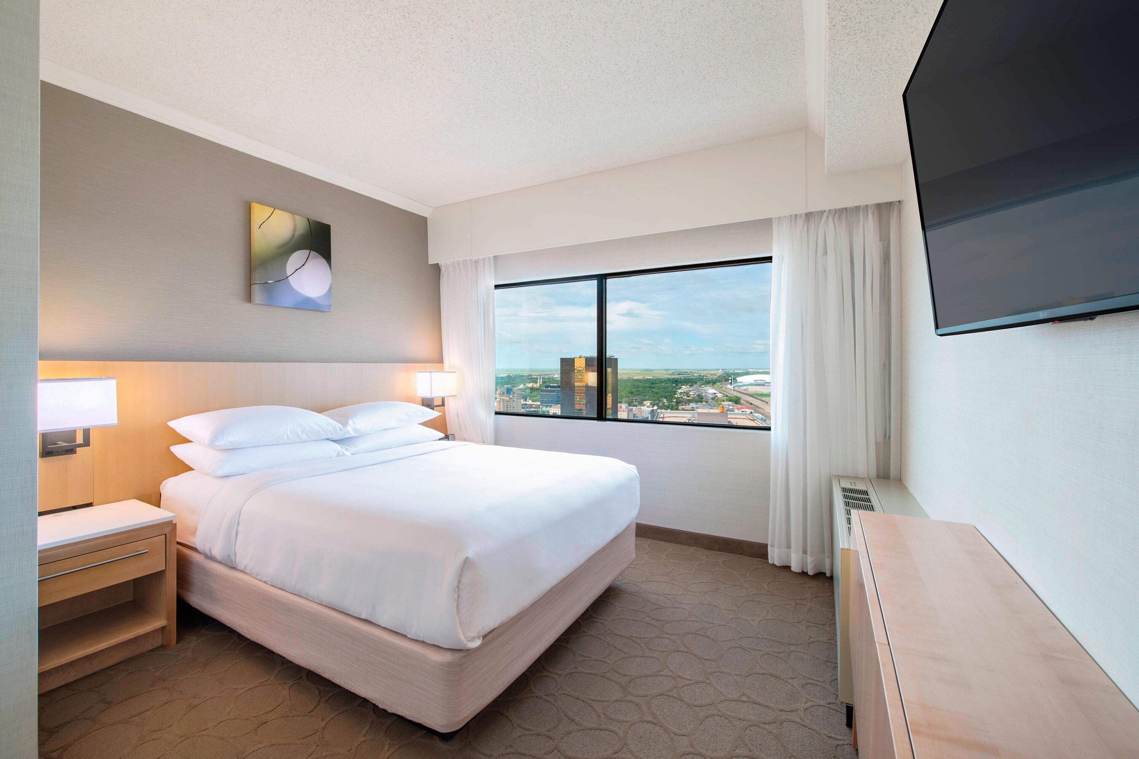 A hotel room with a large bed, a desk, a chair, a TV, and a window with a city view.