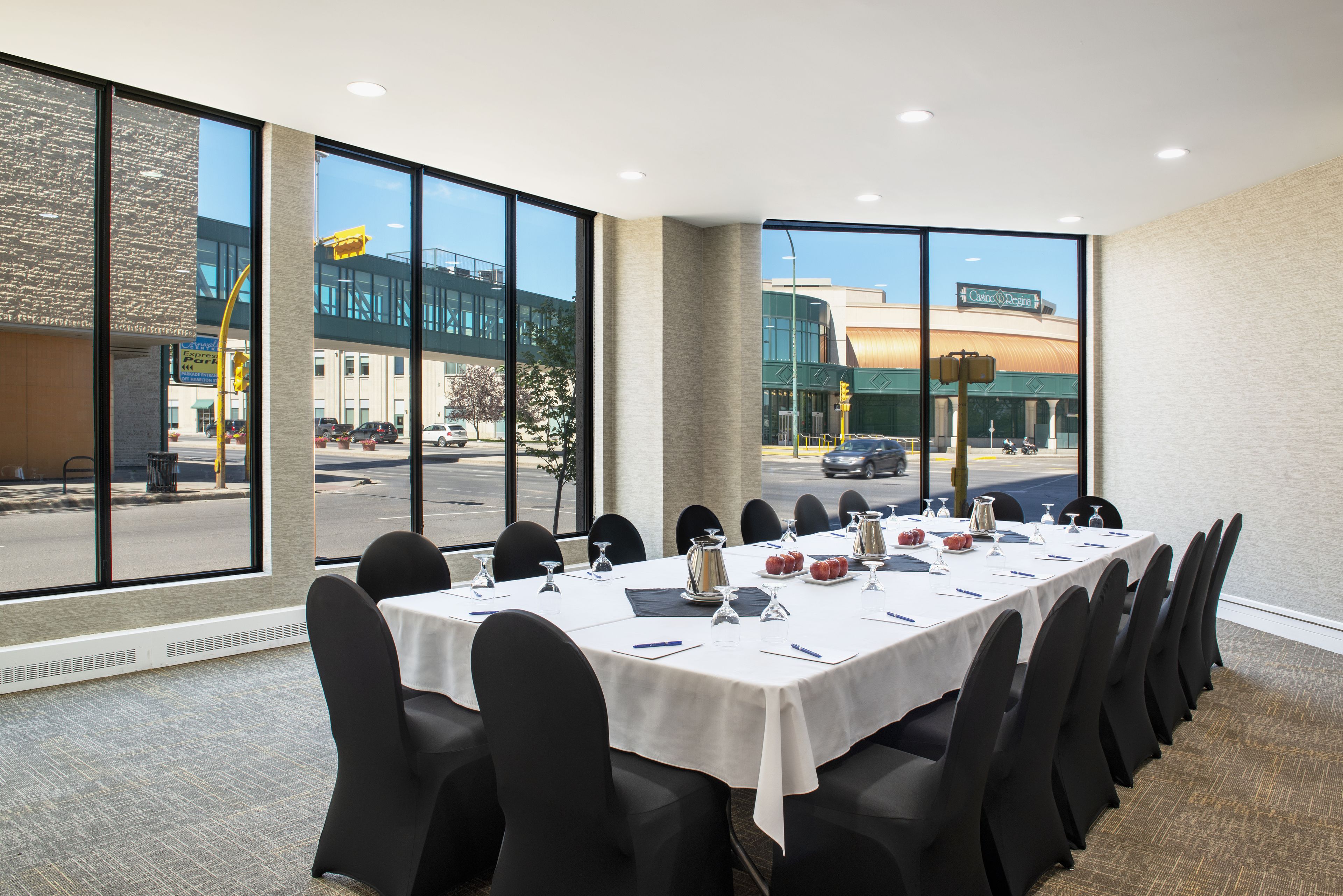 Un salón de conferencias con una larga mesa, sillas negras y ventanas amplias que dan vista a la calle y edificios.