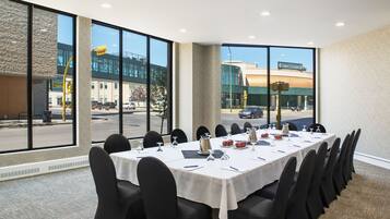Un salón de conferencias con una larga mesa, sillas negras y ventanas amplias que dan vista a la calle y edificios.