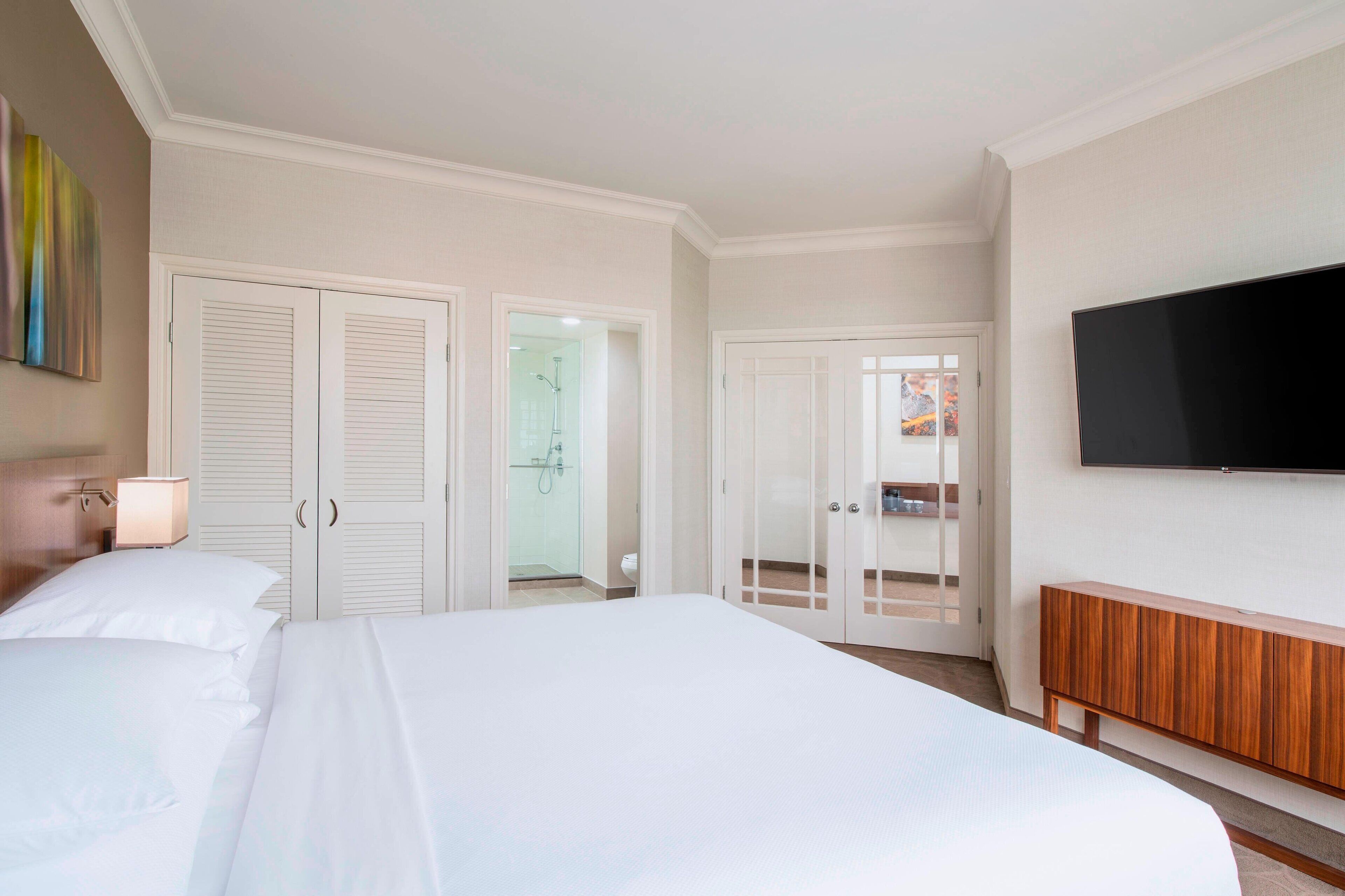 A hotel room with a large bed, a TV mounted on the wall, a wooden cabinet, and a bathroom visible through an open door.
