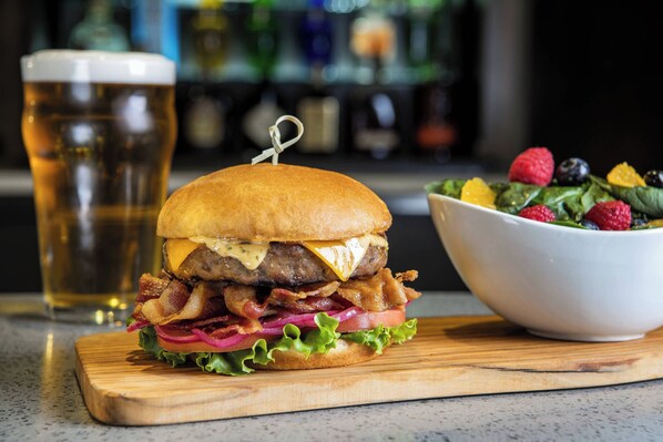 A burger with bacon, cheese, and lettuce on a wooden board, a glass of beer, and a bowl of mixed fruit salad. - Delta Hotels by Marriott Regina (Regina)
