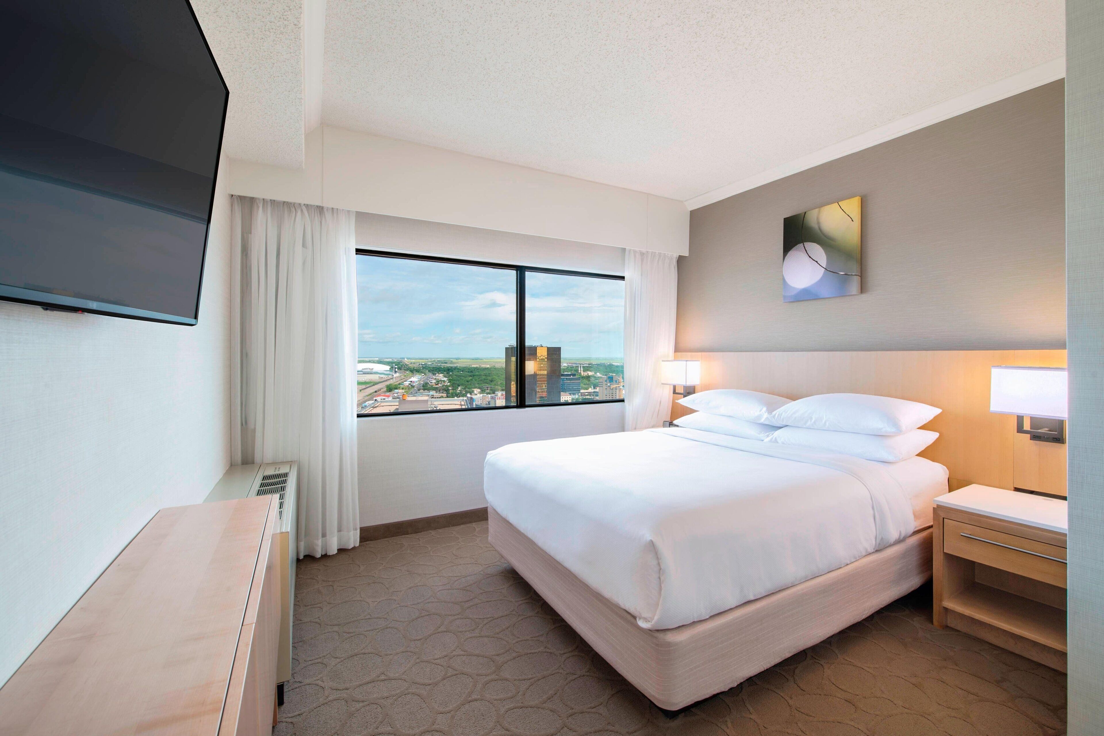 Una habitación de hotel con una cama grande, un televisor de pantalla plana montado en la pared, un ventanal con vistas a la ciudad y una mesita de noche con lámpara.
