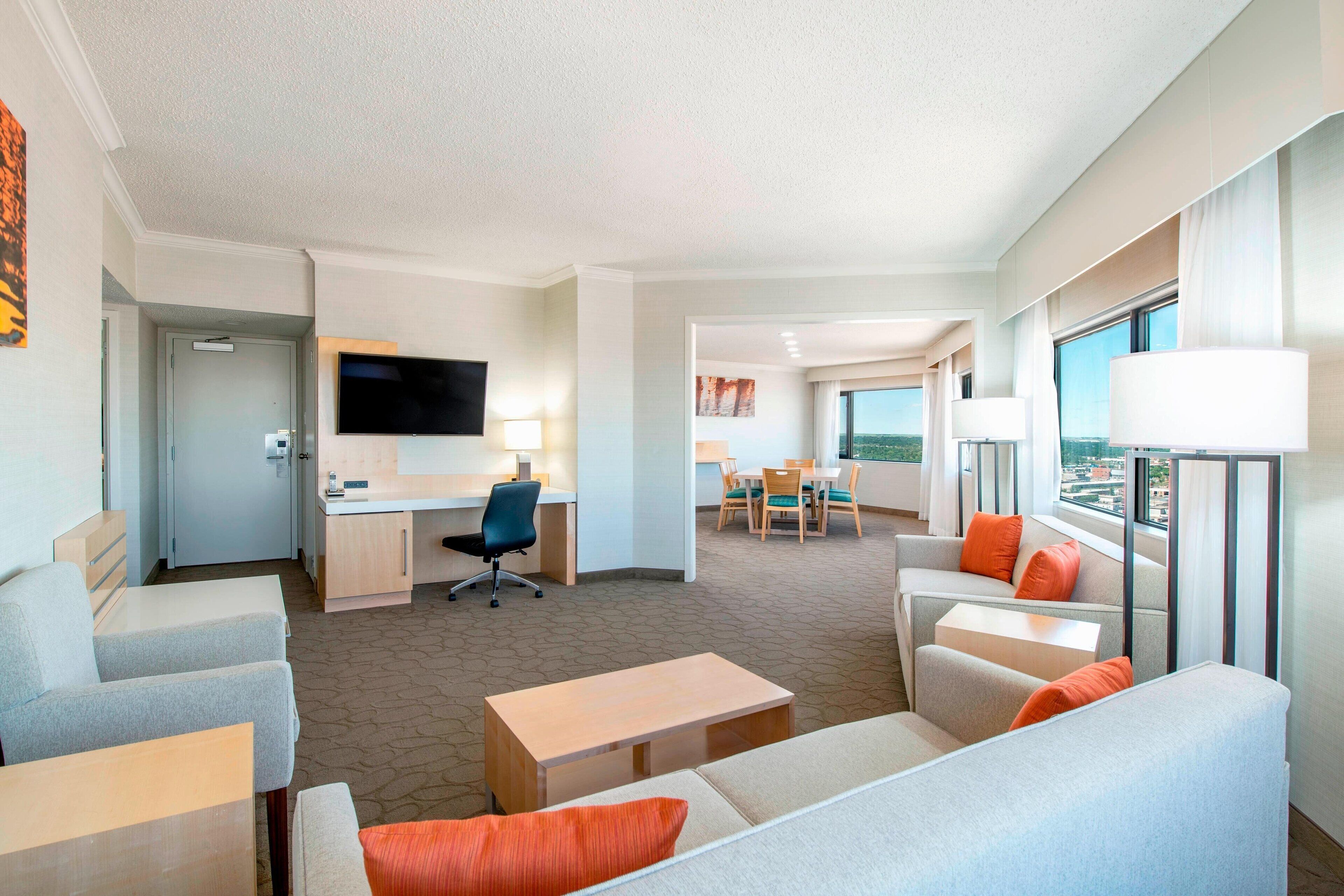 A modern hotel room with a living area, a desk, a flat-screen TV, and a dining area with a view.