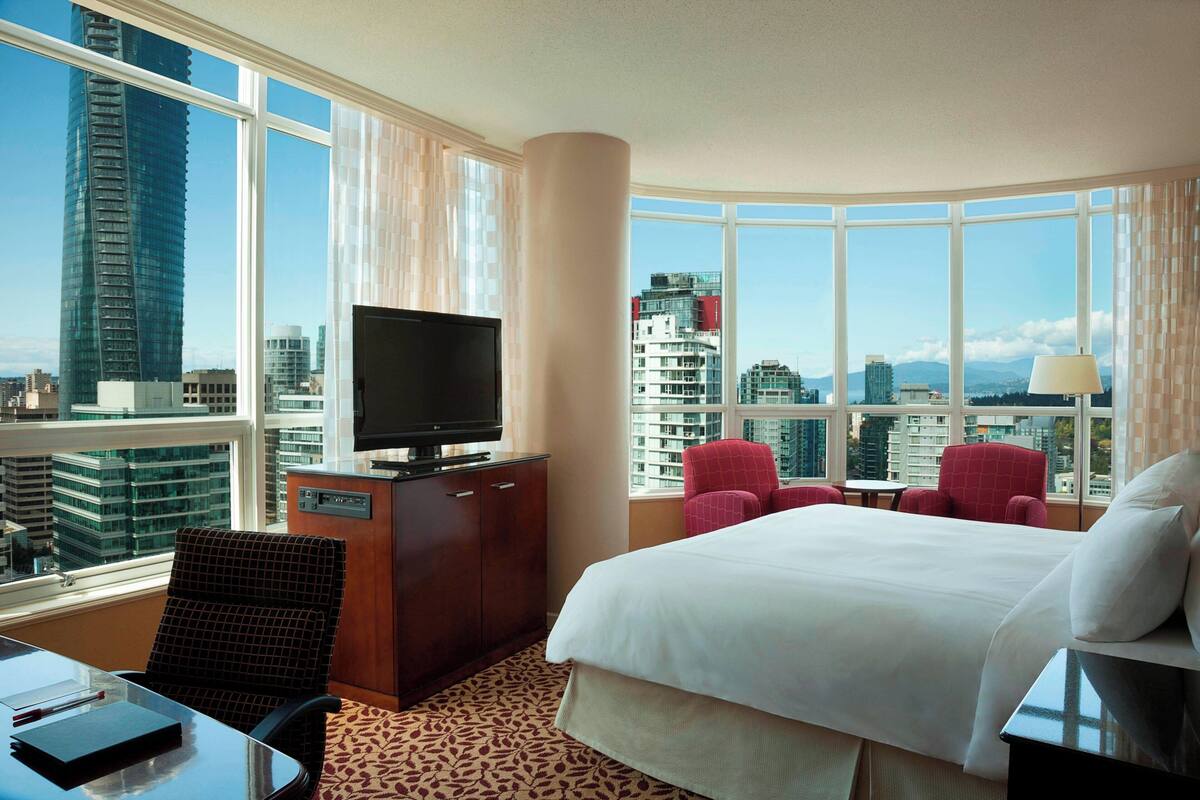 A hotel room with a large bed, a TV on a wooden cabinet, a desk with a chair, and a view of the cityscape through large windows.