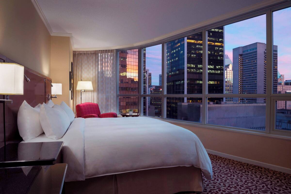 A hotel room with a large bed, a desk, and a chair. The room has a large window with a city view.