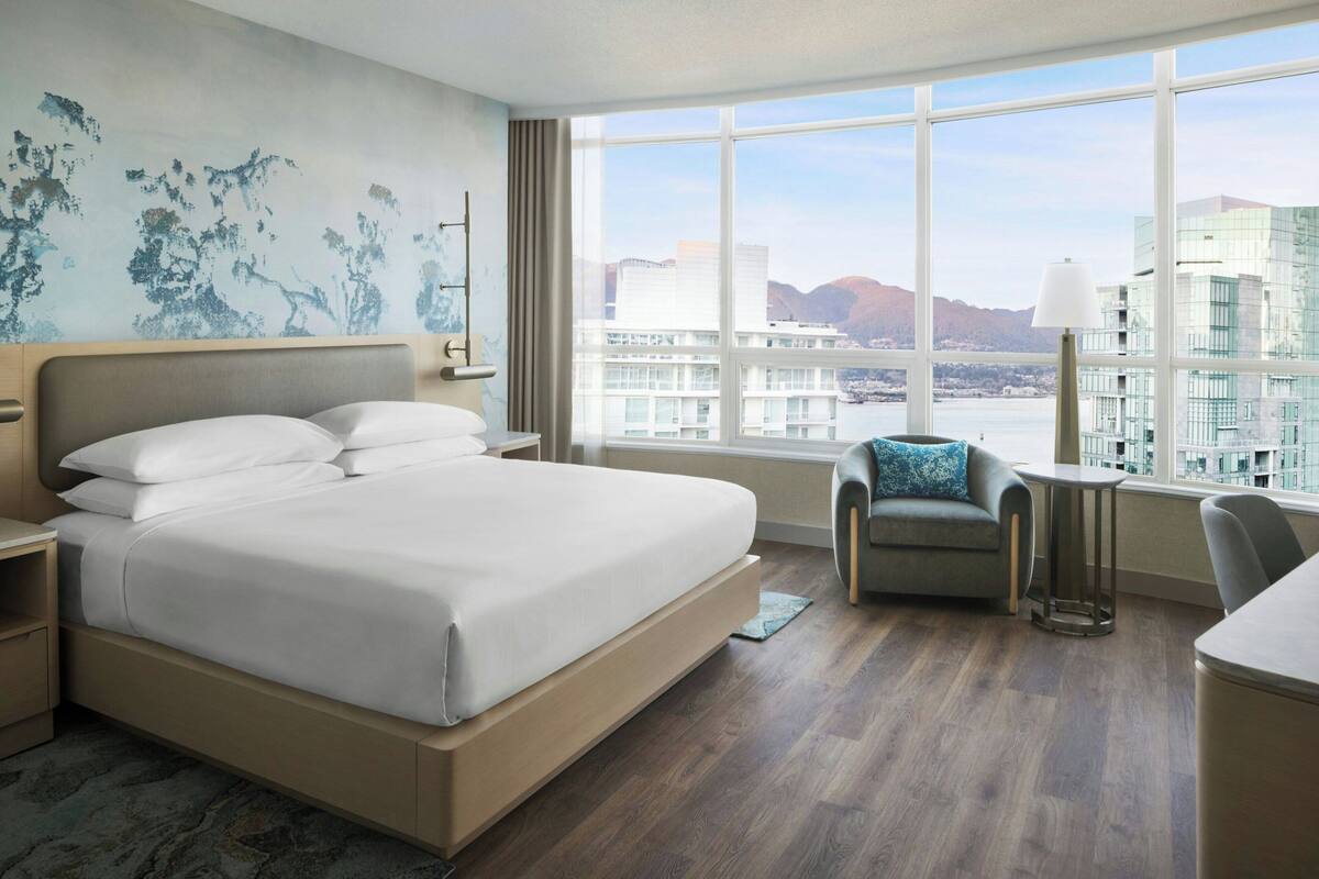 A hotel room with a large bed, a sofa, a small table, and a view of buildings and mountains.