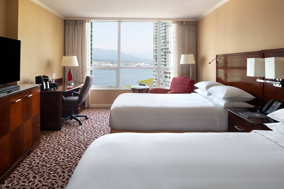 A hotel room with two beds, a desk, a chair, a TV, and a view of a city and mountains.