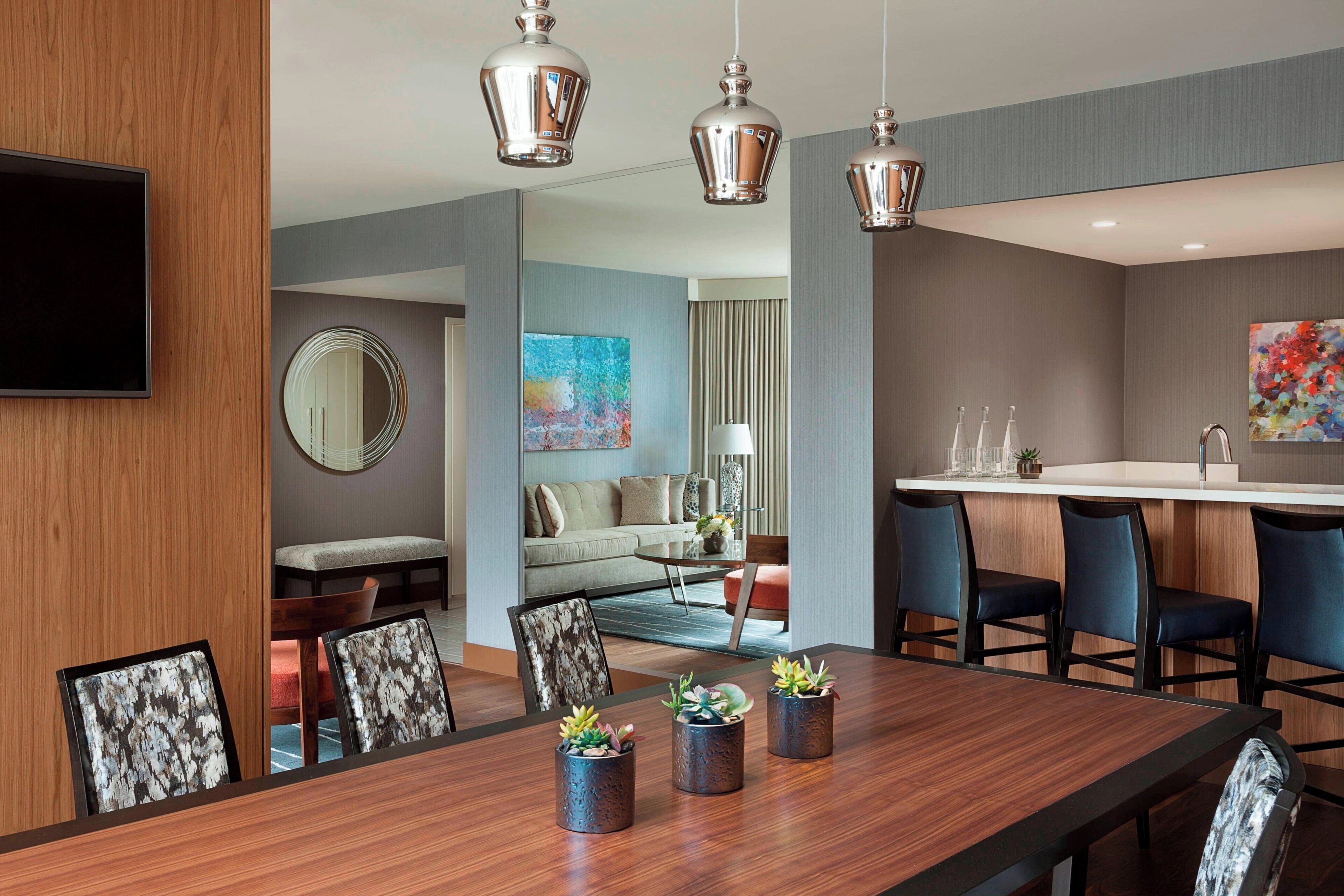 A modern hotel room with a wooden dining table, chairs, and potted plants. There is a large mirror, a sofa, and a painting on the wall.