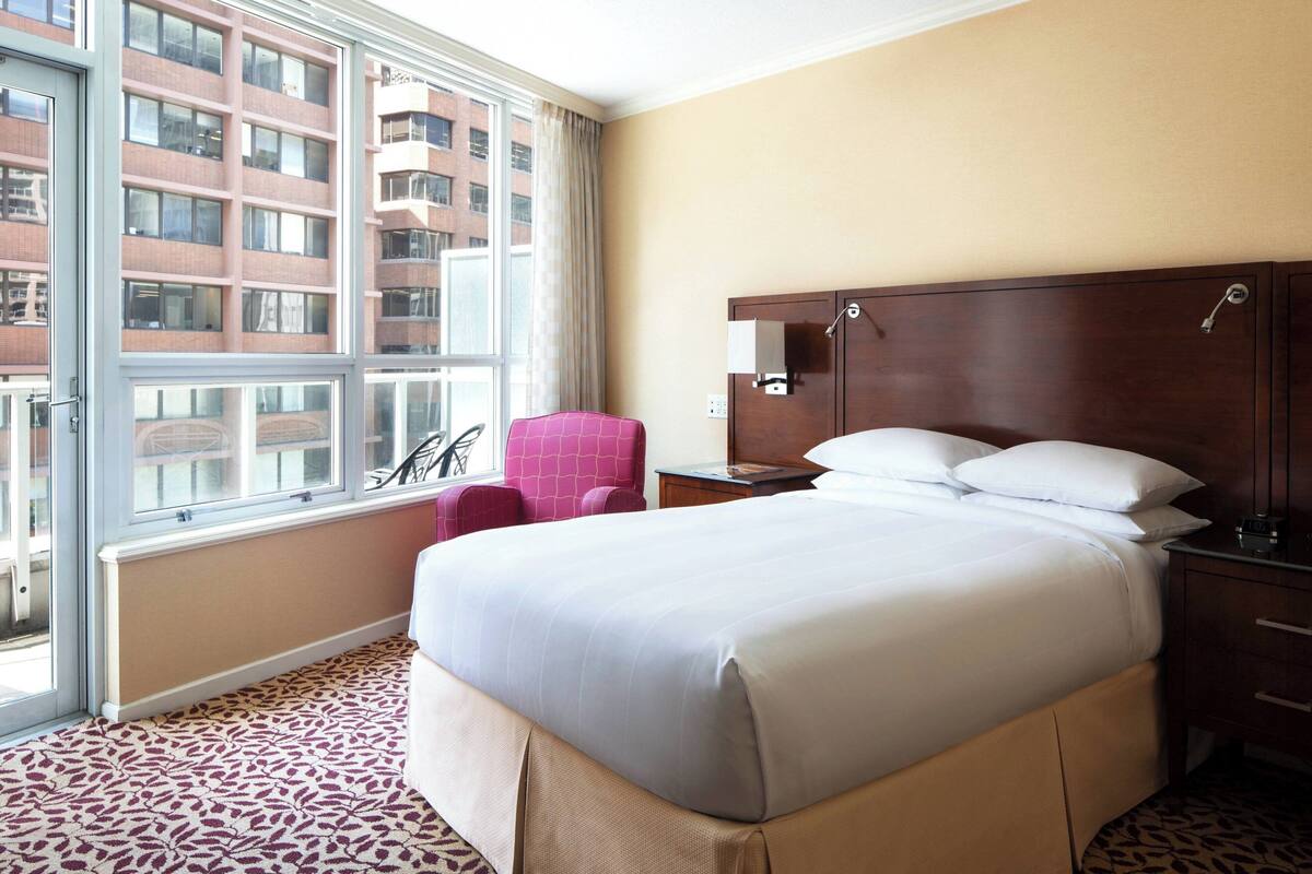 A hotel room with a large bed, a chair, a nightstand, and a window with a view of a building.