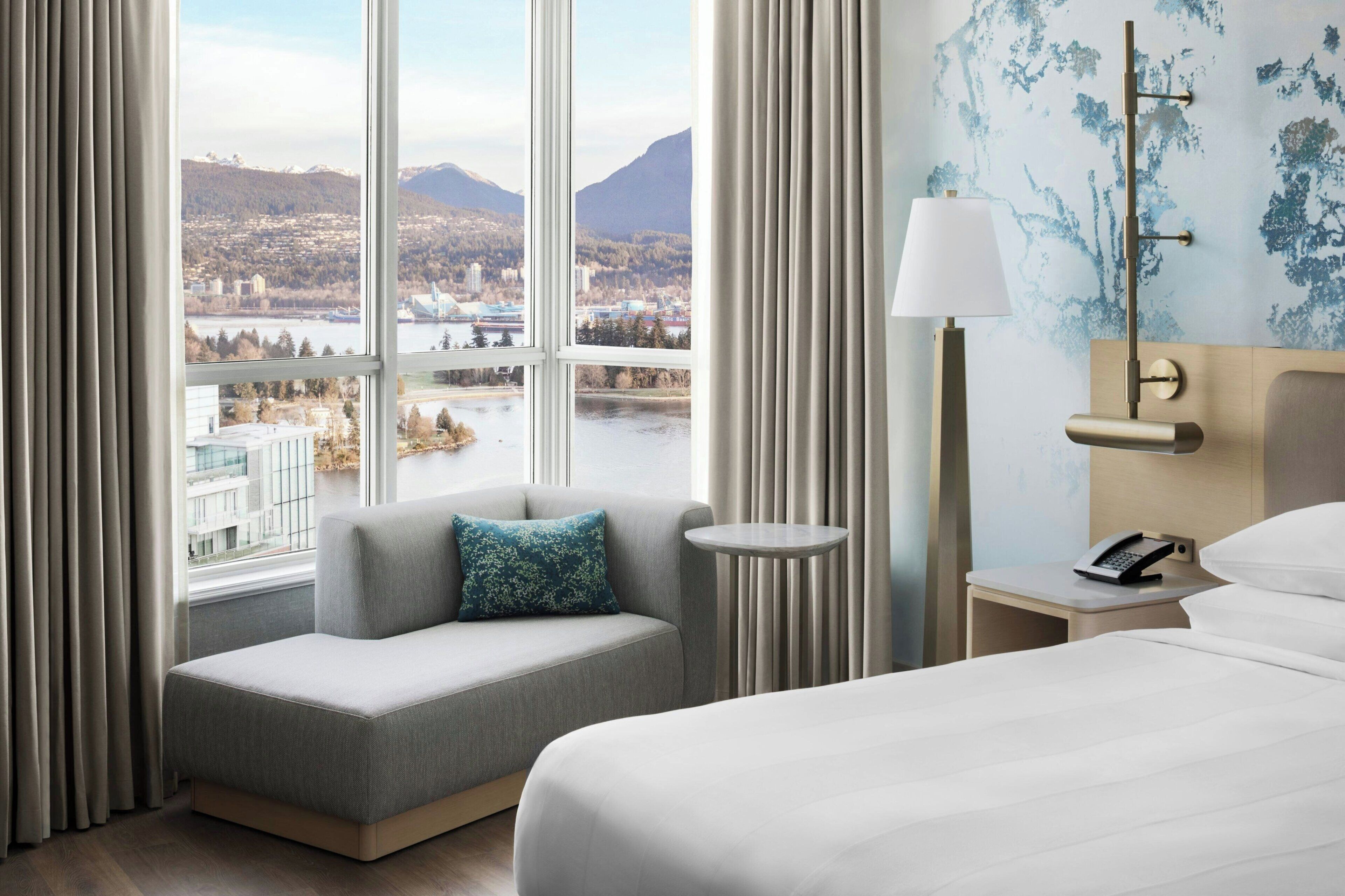 Een hotelkamer met een bank, een bed, een nachtkastje met lamp, een telefoon en uitzicht op een stad en bergen.