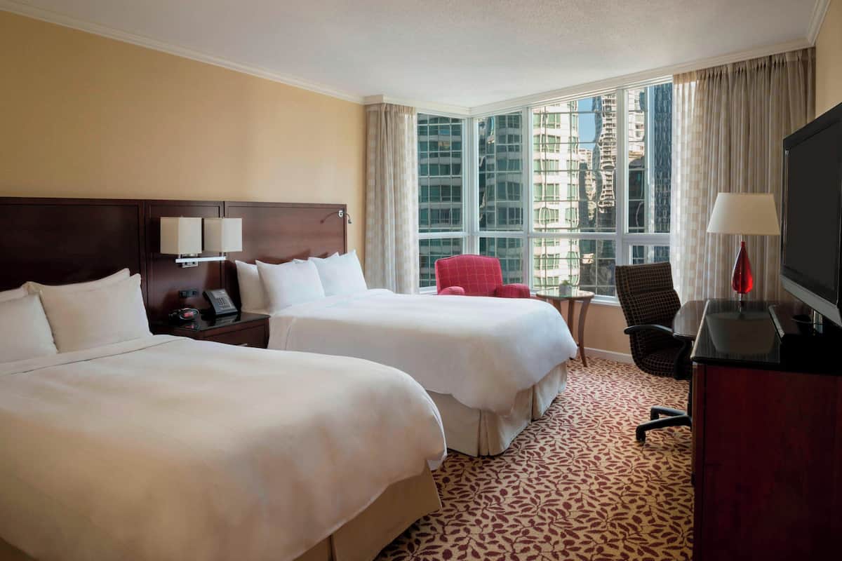 A hotel room with two beds, a desk with a TV, a chair, and a window with a city view.