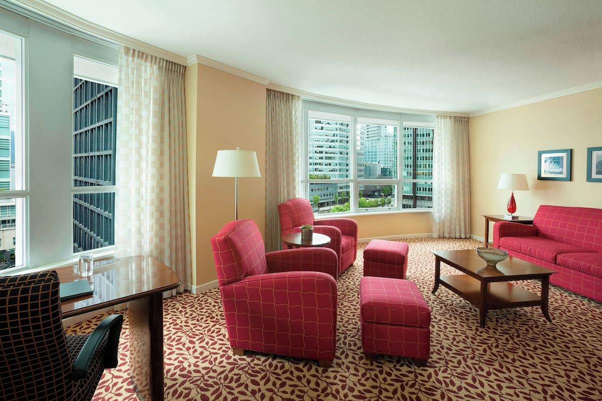 A hotel room with a large window, red upholstered furniture, a patterned carpet, and a city view.