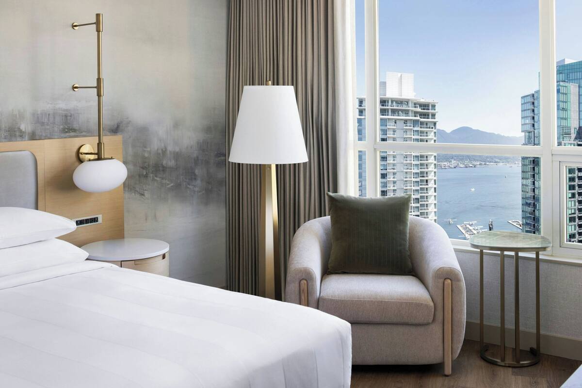 A hotel room with a bed, a chair, a lamp, a small table, and a view of the city and water.