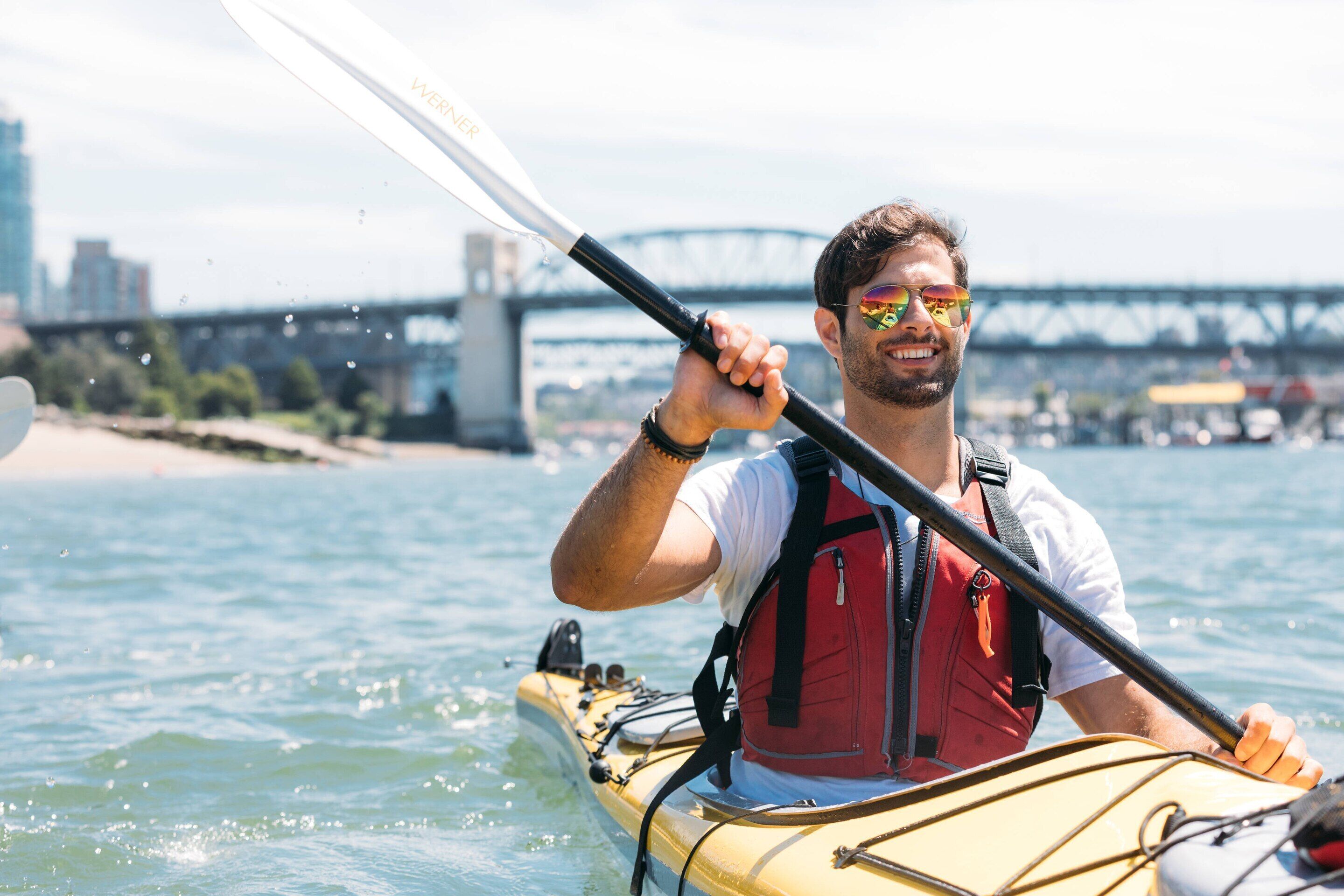 A person kayaking with a paddle, wearing a life jacket, and a cityscape with a bridge in the background.