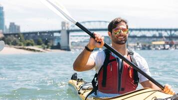 A person kayaking with a paddle, wearing a life jacket, and a cityscape with a bridge in the background.