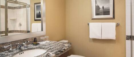 A bathroom with a granite countertop sink, a white toilet, a glass shower enclosure, and a framed picture on the wall.