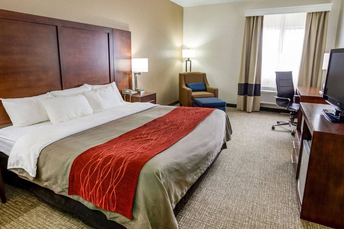 A hotel room with a large bed, a desk with a computer, a chair, a lamp, and a window with curtains.