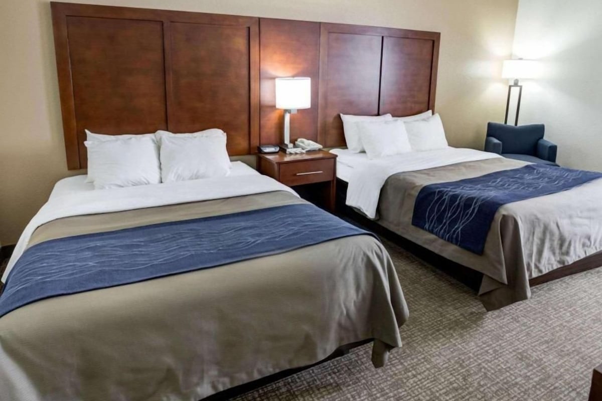 A hotel room with two beds, wooden headboards, bedside tables with lamps, and a carpeted floor.