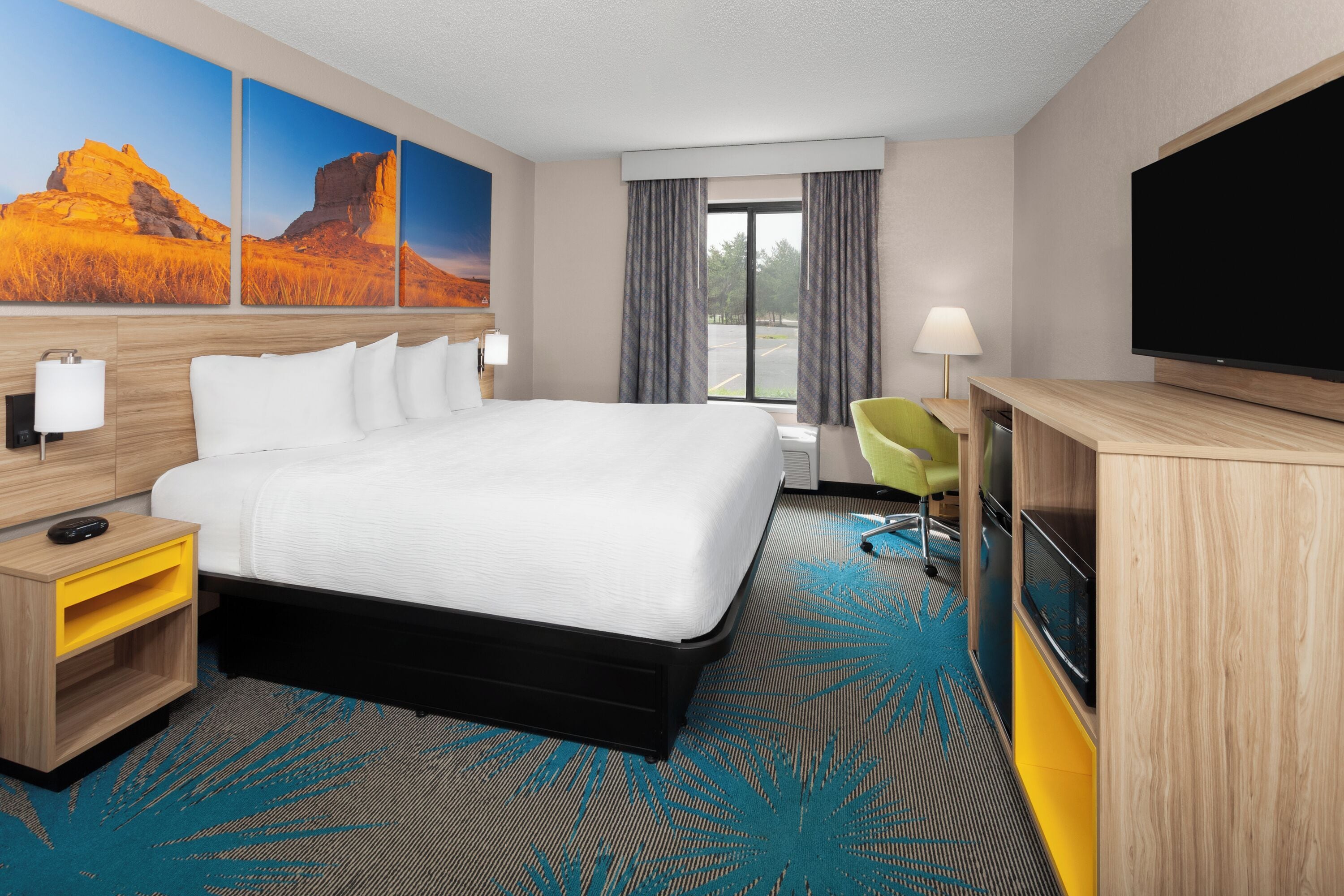 A hotel room with a large bed, a desk with a flat-screen TV, a green chair, and a window with curtains.