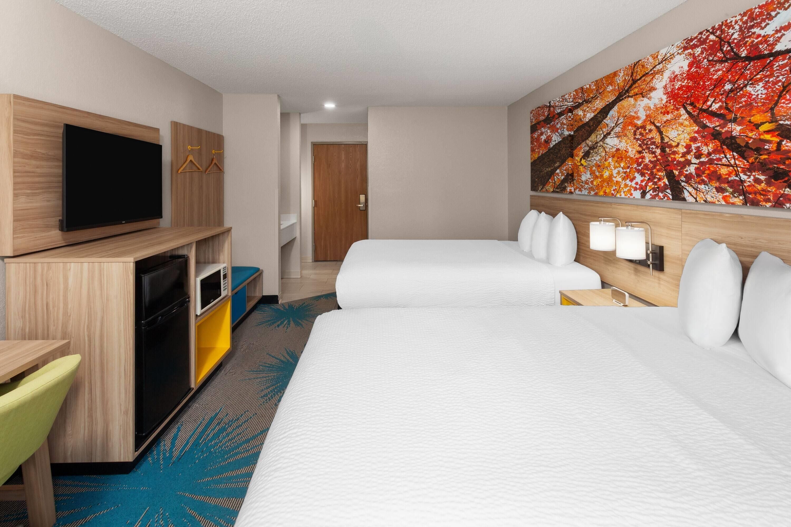A hotel room with two beds, a TV, a desk with a microwave, and a painting of autumn leaves.