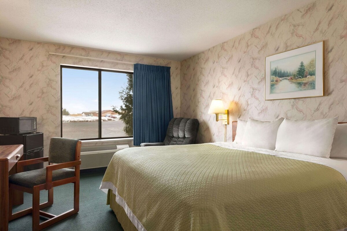 Una habitación de hotel con cama, escritorio y silla, televisión, ventana con vista al exterior y un cuadro en la pared.