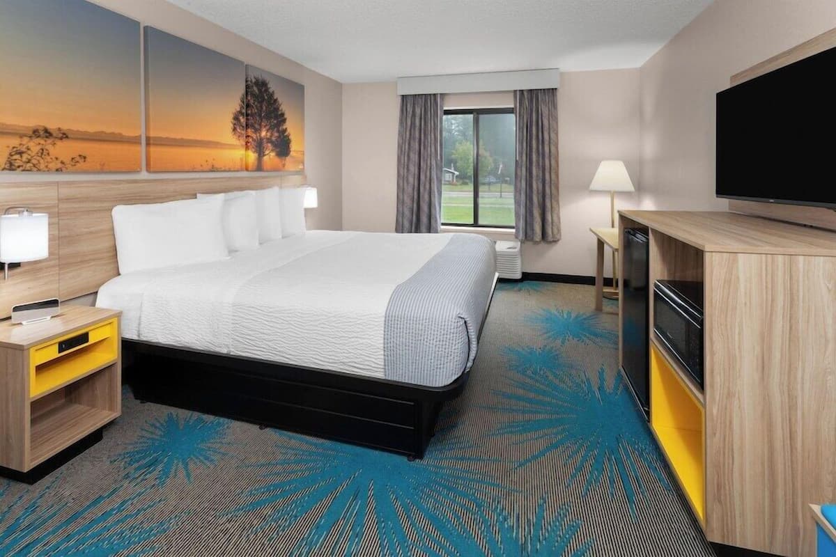 Habitación de hotel con cama, televisor de pantalla plana, escritorio con lámpara y ventana grande con cortinas.