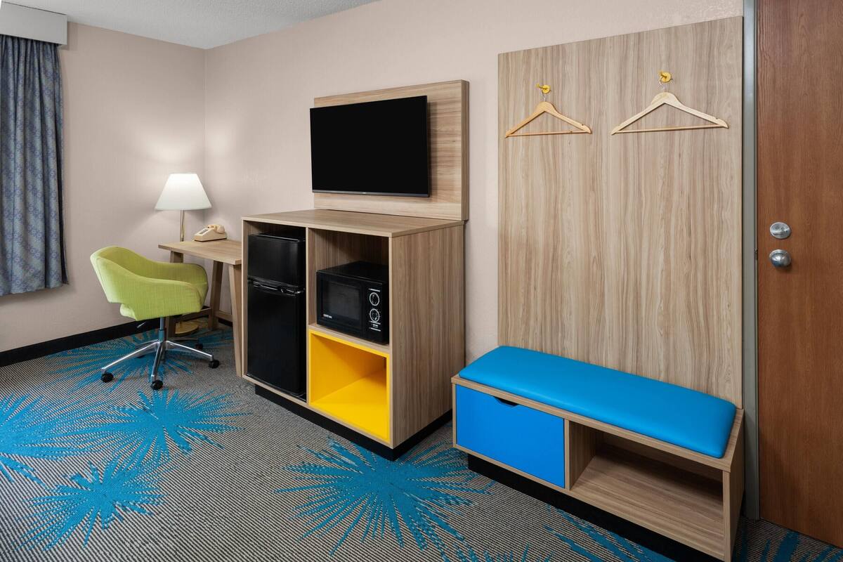 Habitación de hotel con escritorio, silla, televisor, armario y una banca.