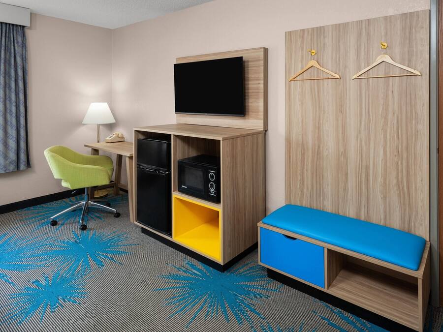 A hotel room with a desk, chair, television, wardrobe, and a bench.