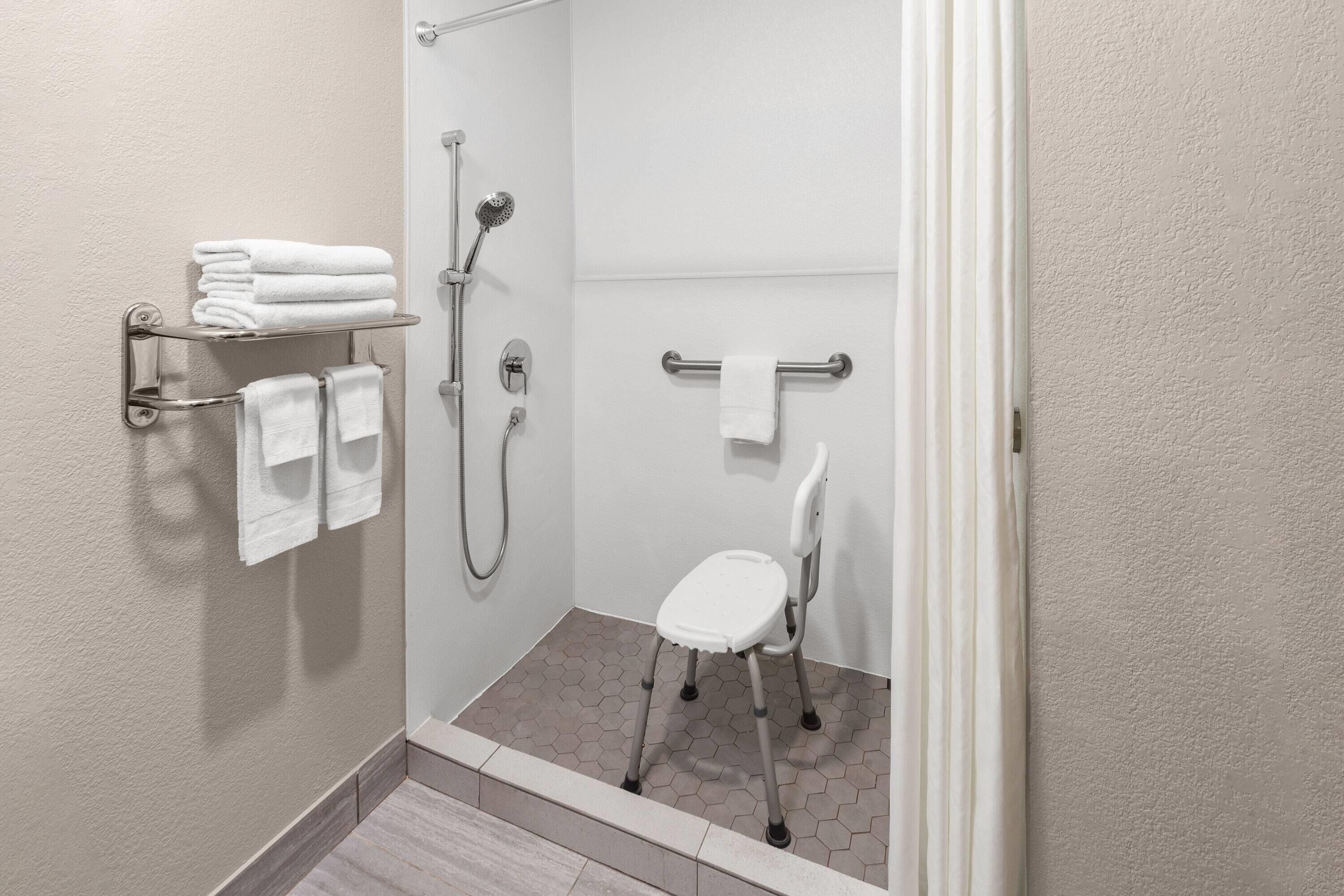 A shower area with a white chair, a towel rack with folded towels, and a showerhead.
