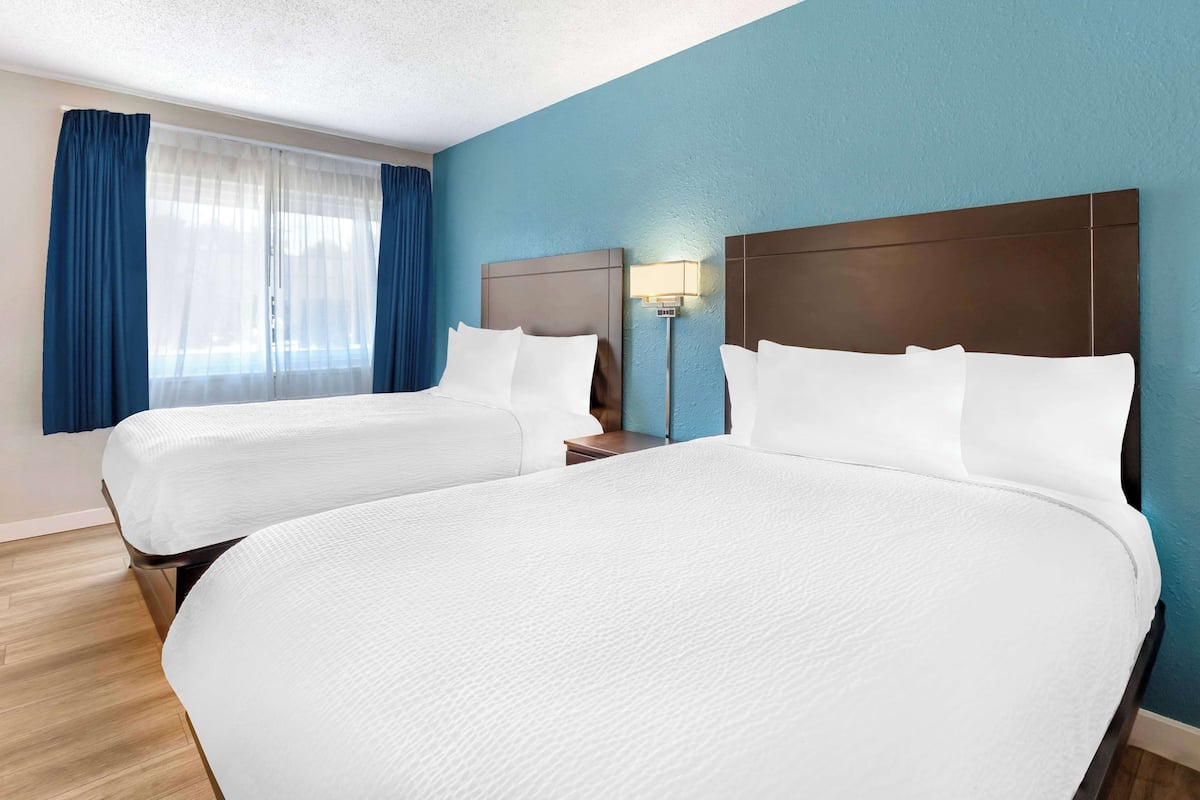 Habitación de hotel con dos camas, una mesita de noche, una lámpara y un ventanal con cortinas azules.