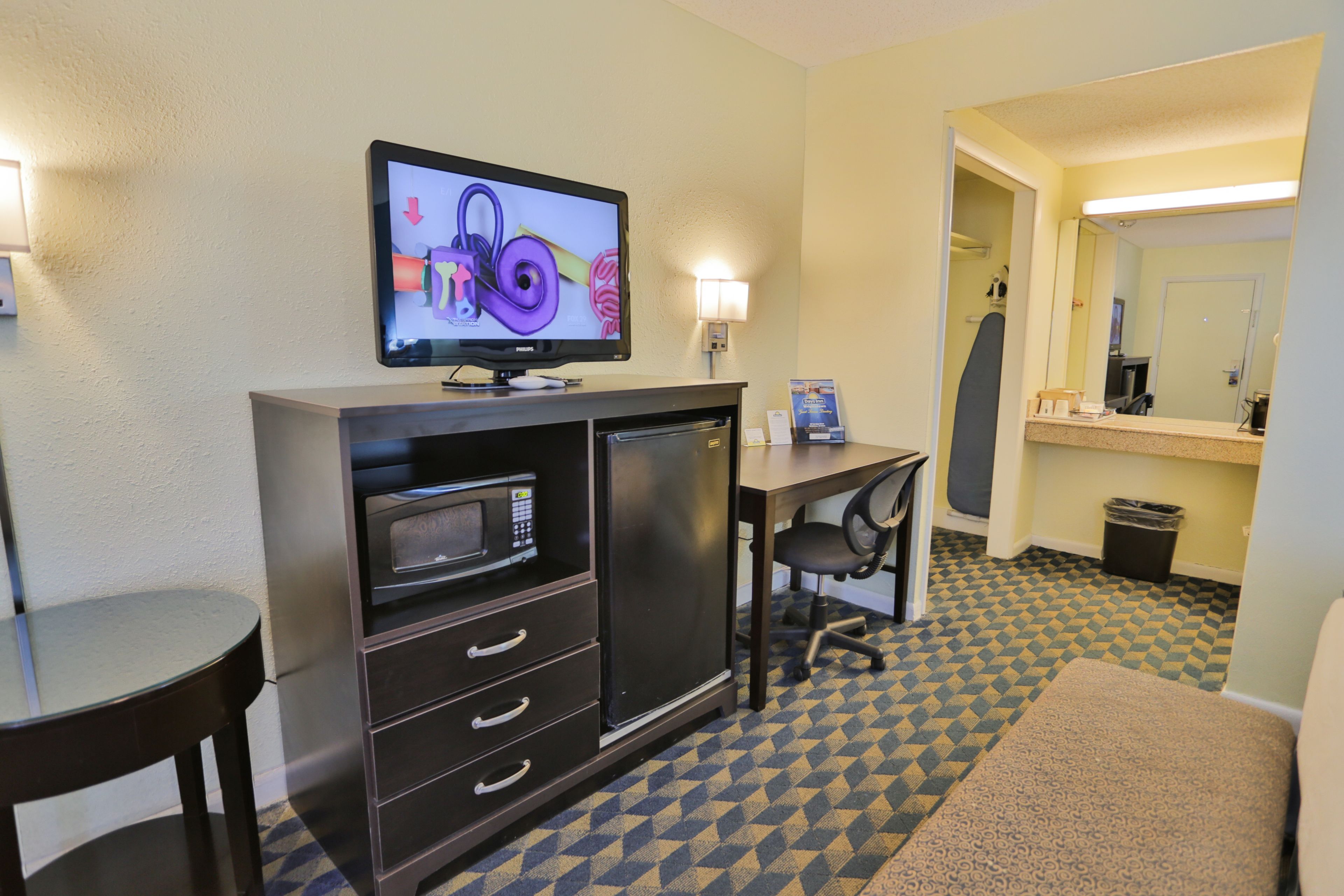 Habitación de hotel con televisión de pantalla plana, microondas, escritorio, silla, mesita y alfombra estampada.