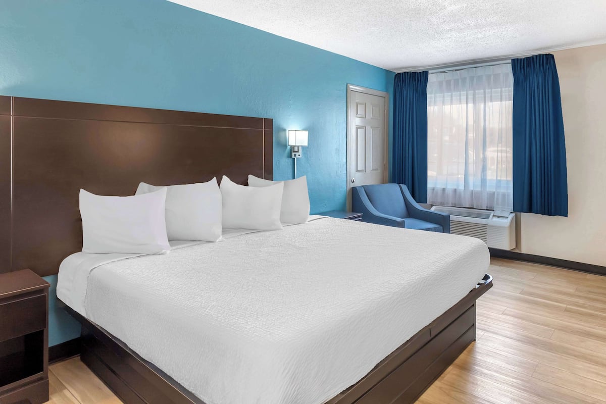 Habitación de hotel con una cama grande, una mesita de noche, un escritorio, una silla, un ventanal con cortinas y un equipo de aire acondicionado.