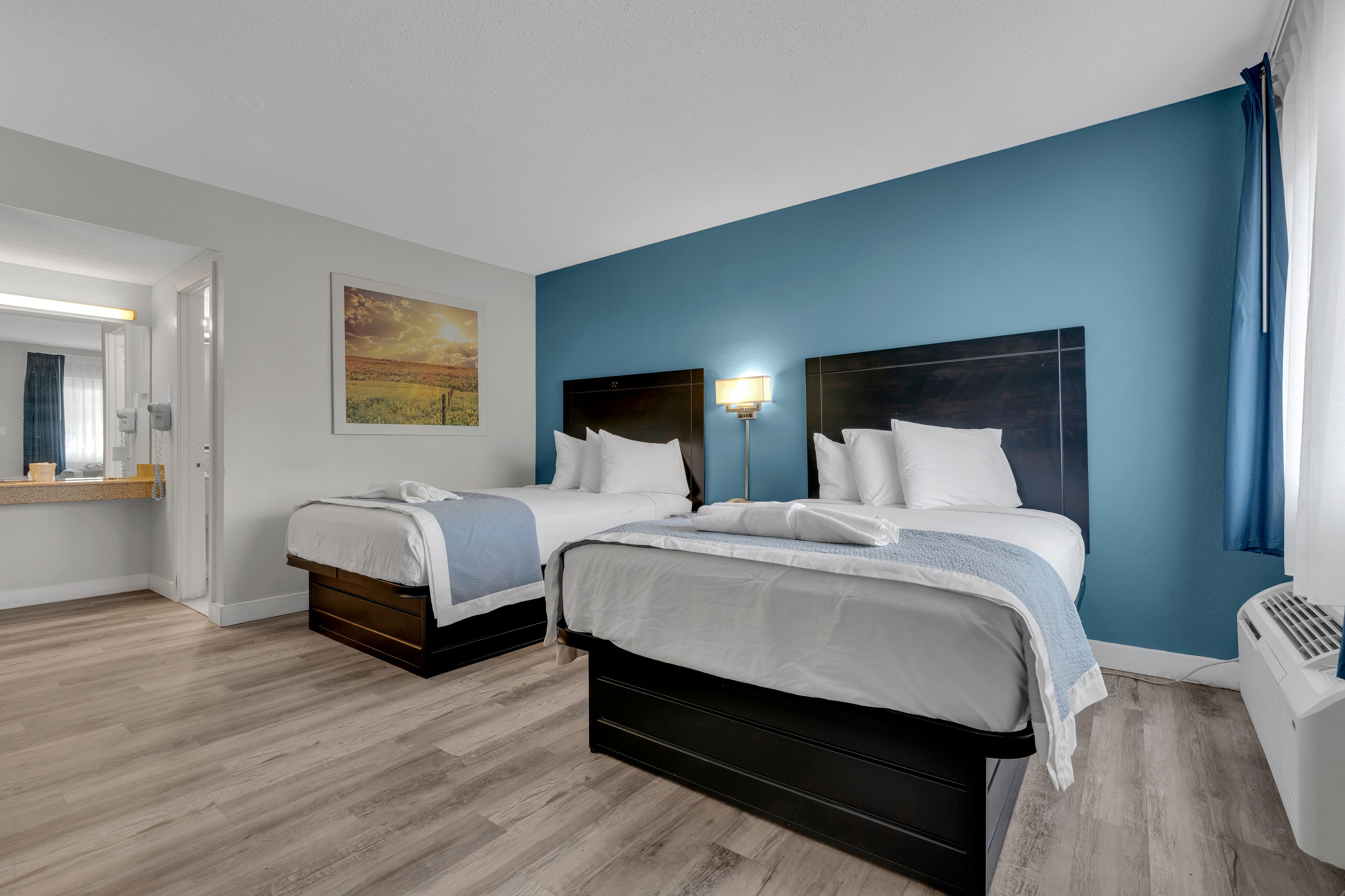 A hotel room with two beds, a large painting on the wall, a bedside table with a lamp, and a window with curtains.