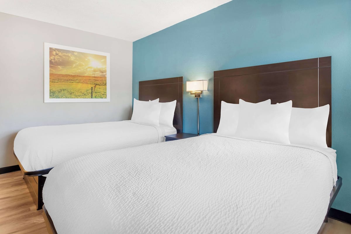 Habitación de hotel con dos camas, una mesita de noche, una lámpara de pared y un cuadro con una imagen de un atardecer.