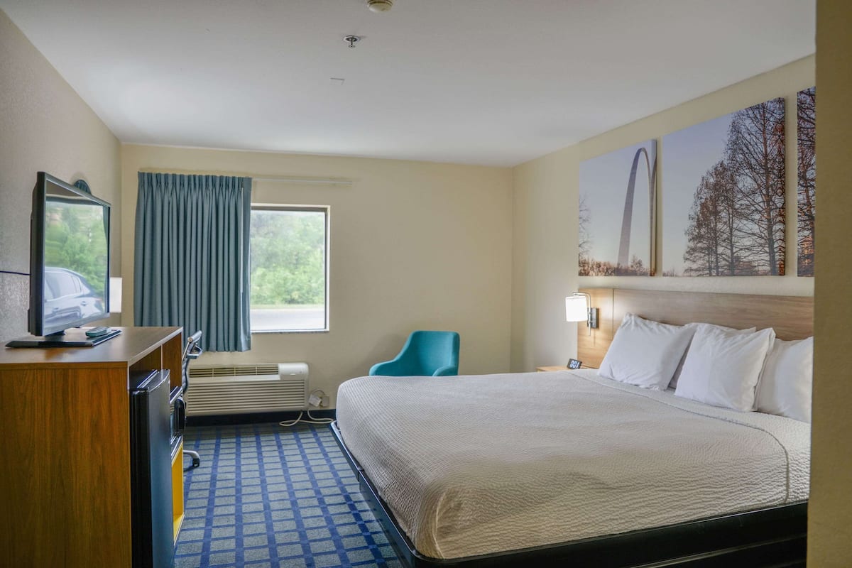 A hotel room with a bed, a desk with a television, a chair, a window with curtains, and a wall with a painting.