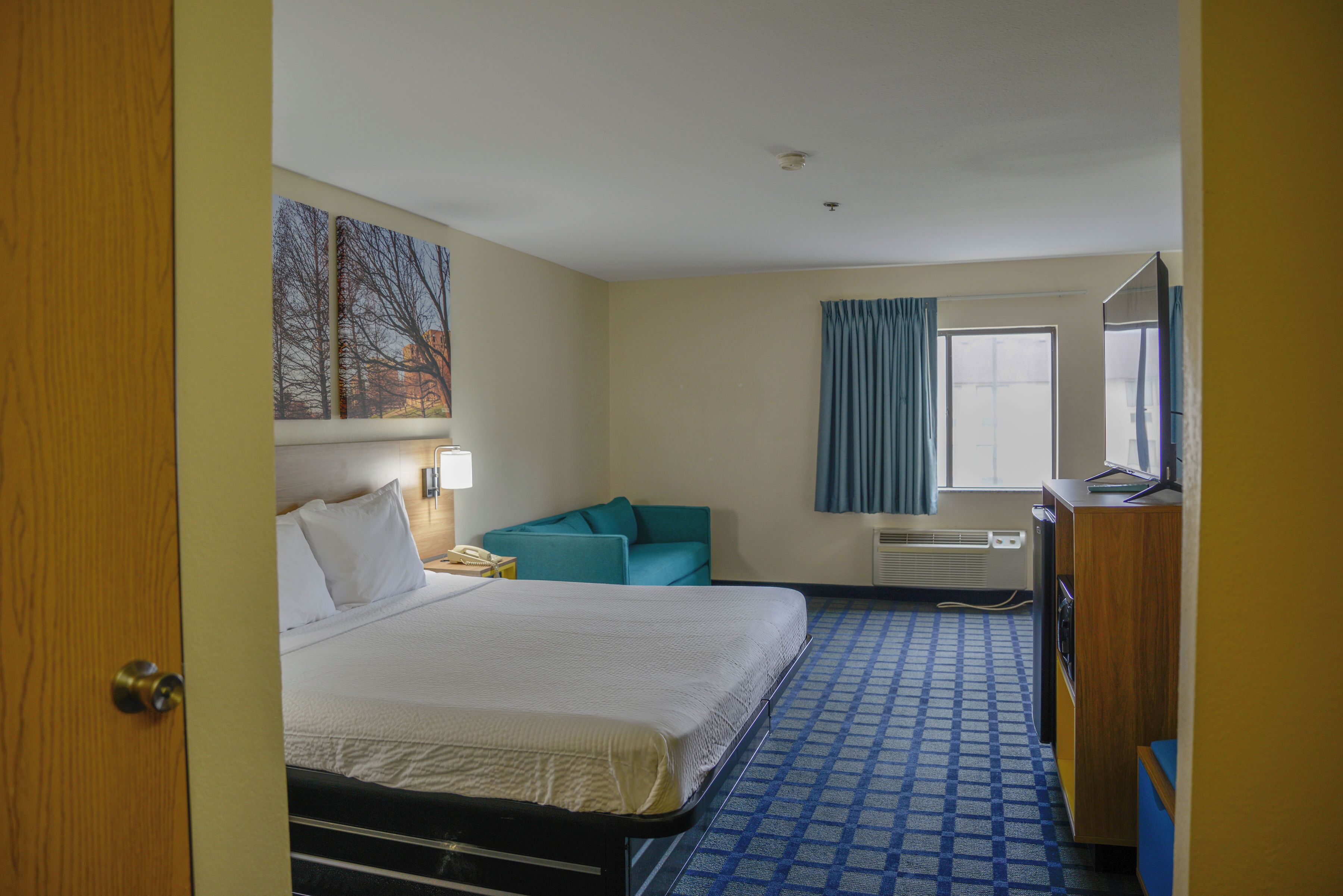 A hotel room with a bed, a desk, a chair, a window with curtains, and a wall-mounted TV.