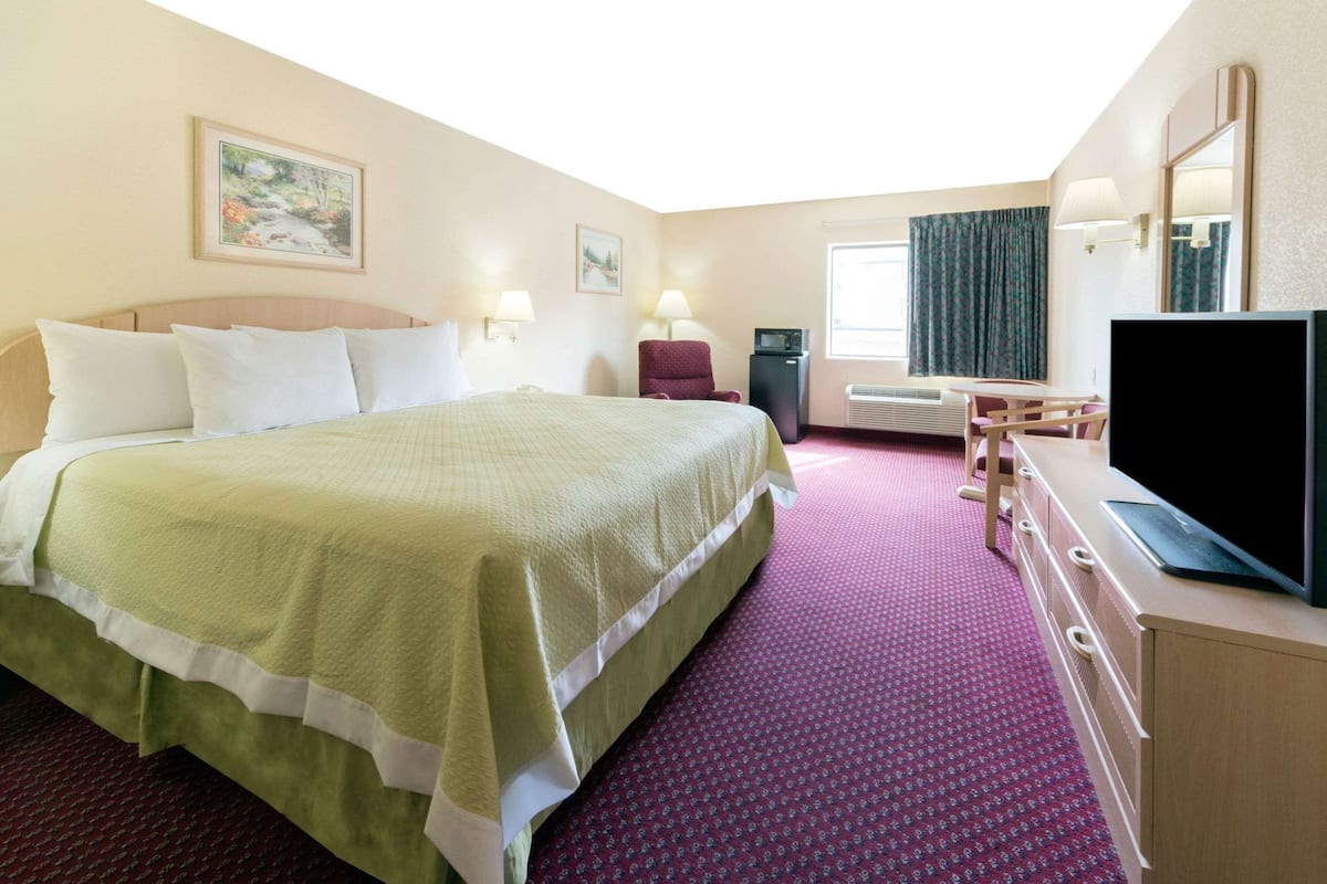 A hotel room with a large bed, a desk with a flat-screen TV, a dresser, and a window with curtains.