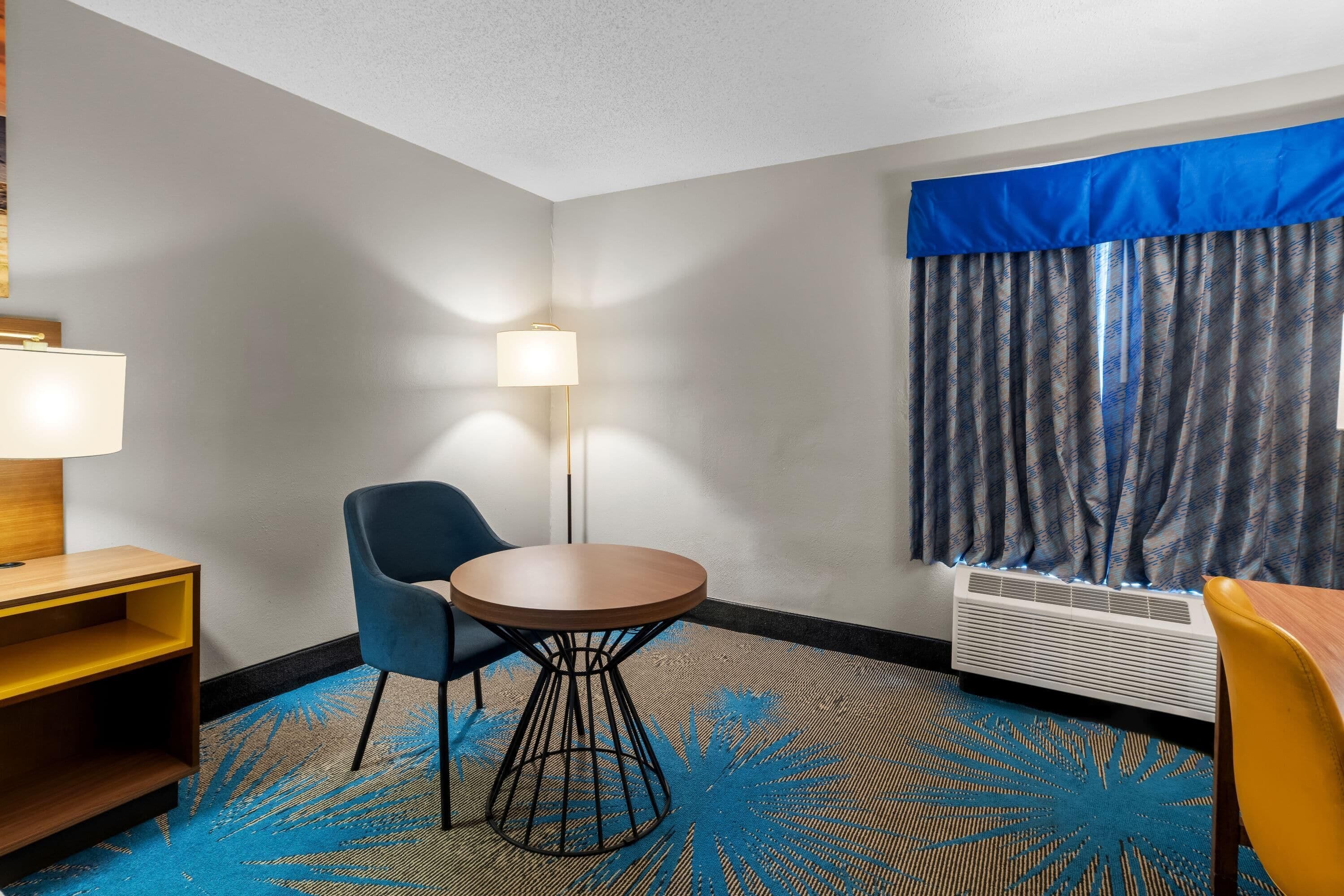 A hotel room with a round table, two chairs, a lamp, a heater, and a window with curtains.
