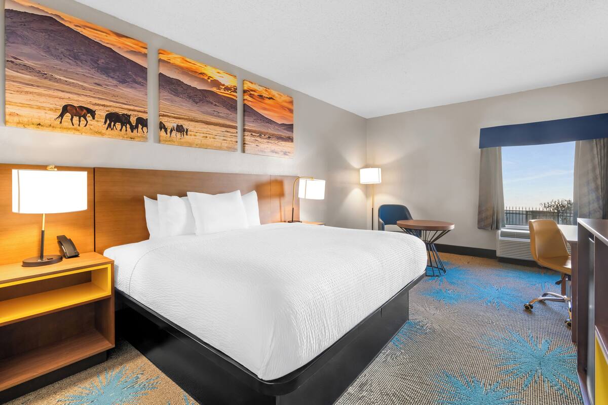A hotel room with a large bed, bedside tables, a desk, and a chair. There are wall-mounted lamps and a window with curtains.