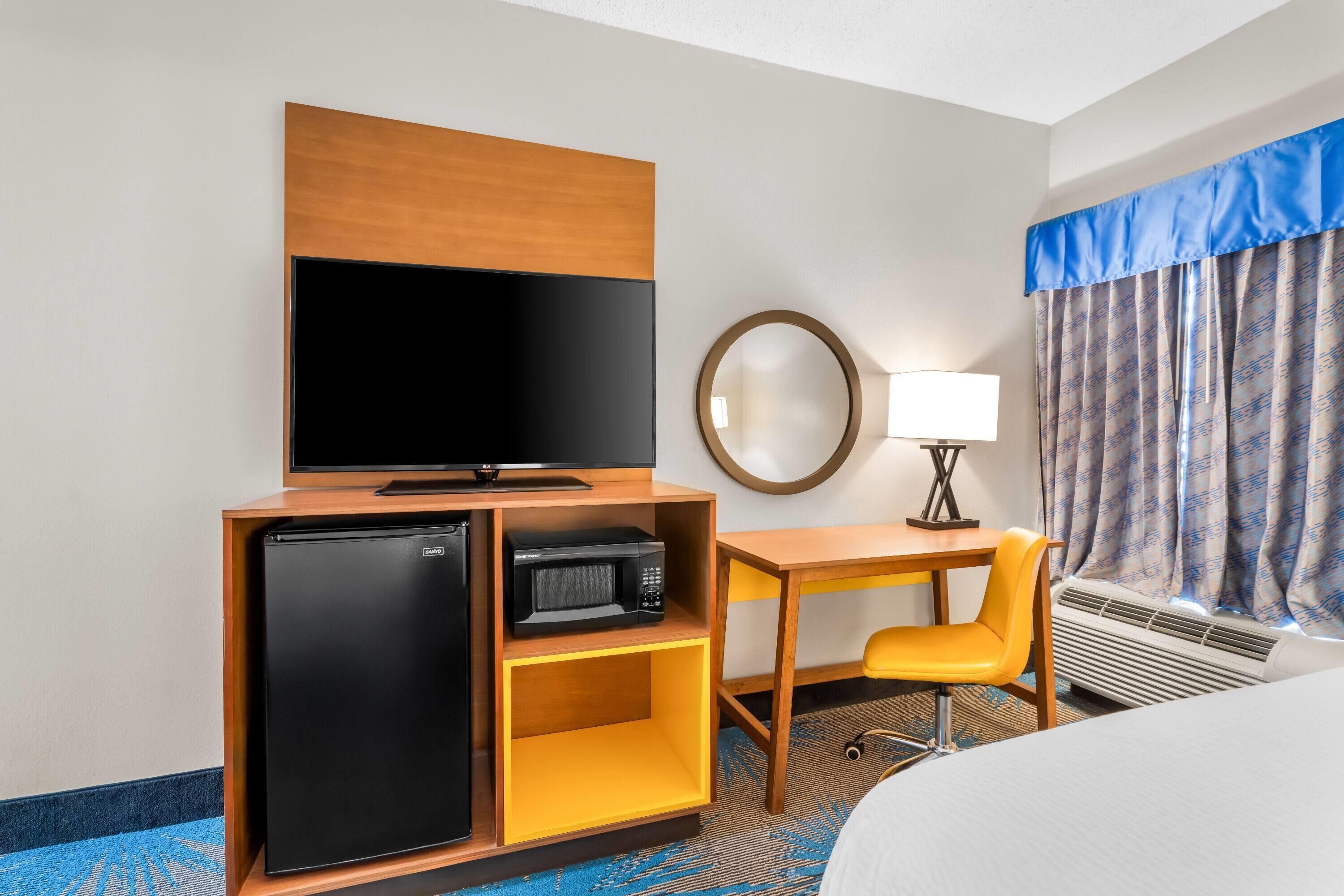 Une chambre d’hôtel équipée d’un lit, d’une télévision, d’un mini-réfrigérateur, d’un four à micro-ondes, d’un bureau avec une chaise, d’une lampe et d’une fenêtre avec des rideaux.