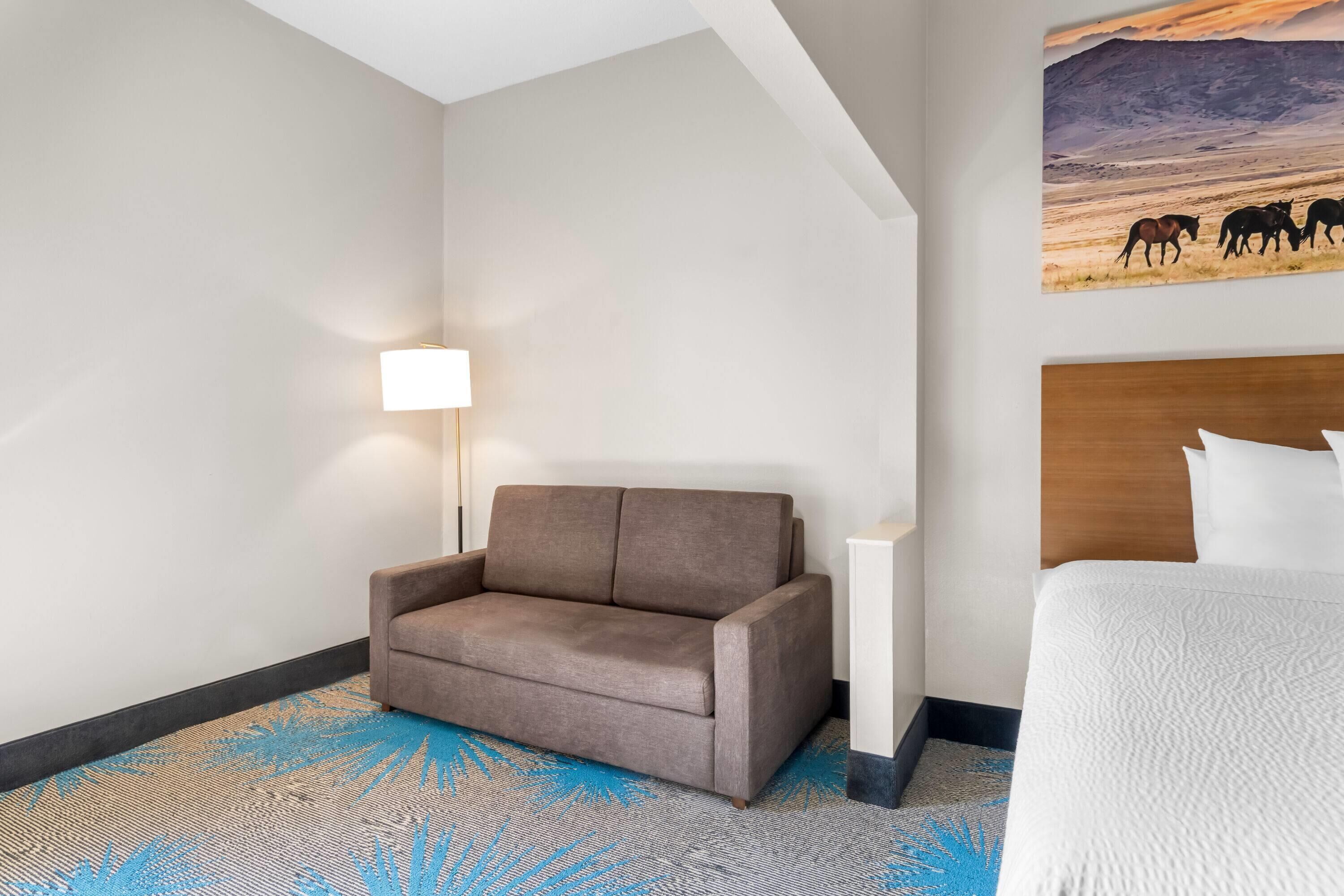 A hotel room with a brown sofa, a white bed, a lamp, and a painting of horses on the wall.