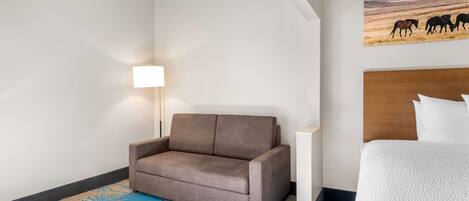 A hotel room with a brown sofa, a white bed, a lamp, and a painting of horses on the wall.