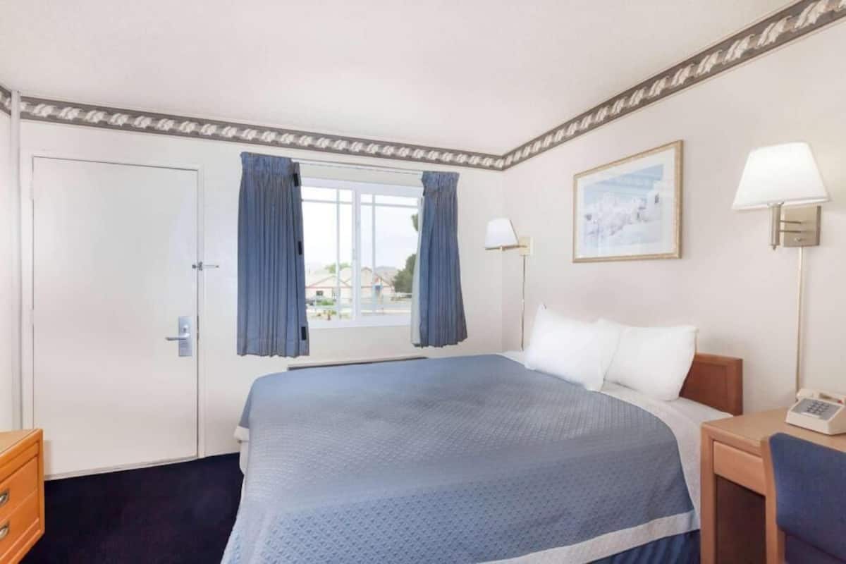 A hotel room with a bed, a desk with a lamp, a window with curtains, a door, and a framed picture on the wall.