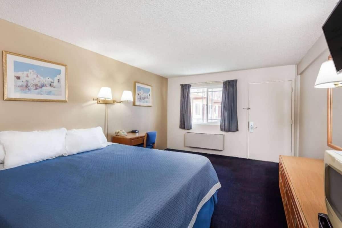 A hotel room with a bed, two wall-mounted lamps, a desk with a chair, a television, and a window with curtains.