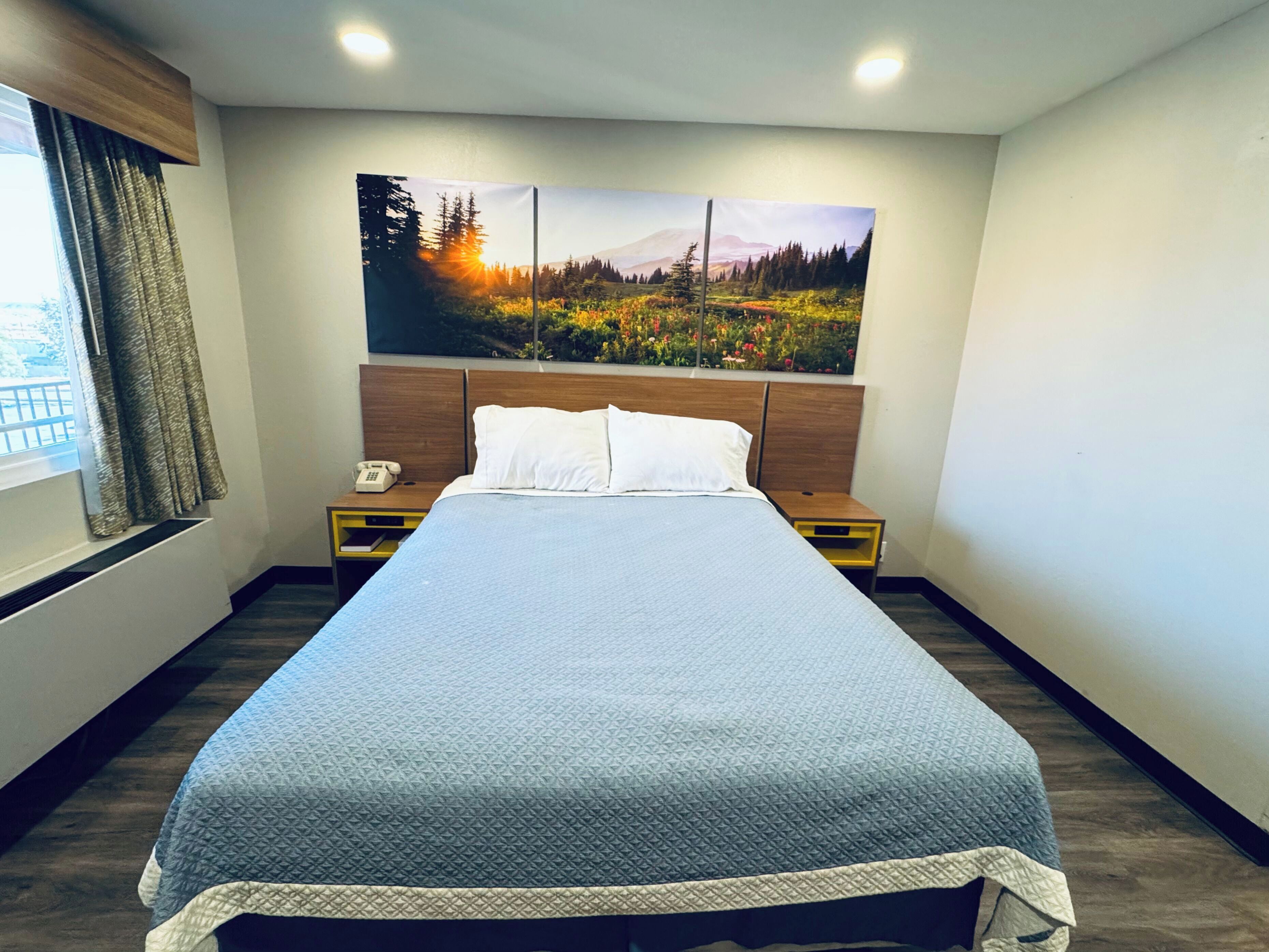 A neatly arranged hotel room with a large bed, a headboard with a painting, a bedside table with a phone, and a window with curtains.