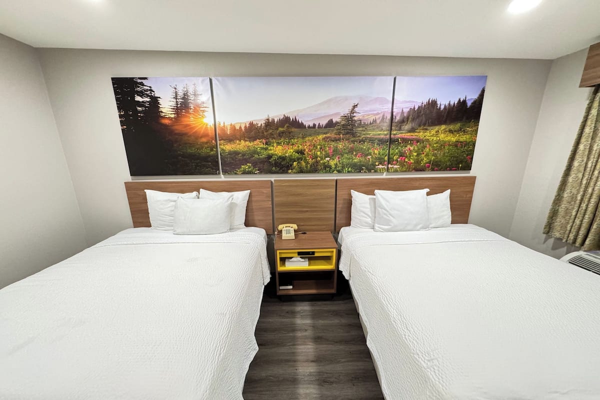 A hotel room with two single beds, a wooden headboard, a nightstand with a telephone, and a large wall mural of a forest landscape.