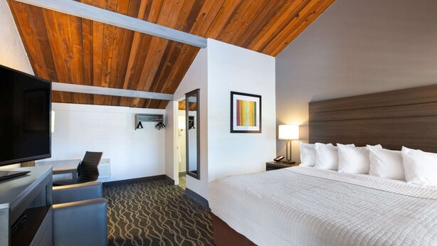 A hotel room with a wooden ceiling, a bed with white bedding, a television, a desk with a chair, and a patterned carpet. - Days Inn by Wyndham Ukiah (Ukiah)