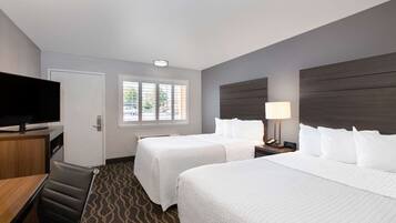 A hotel room with two beds, a TV, a desk, and a window with blinds.