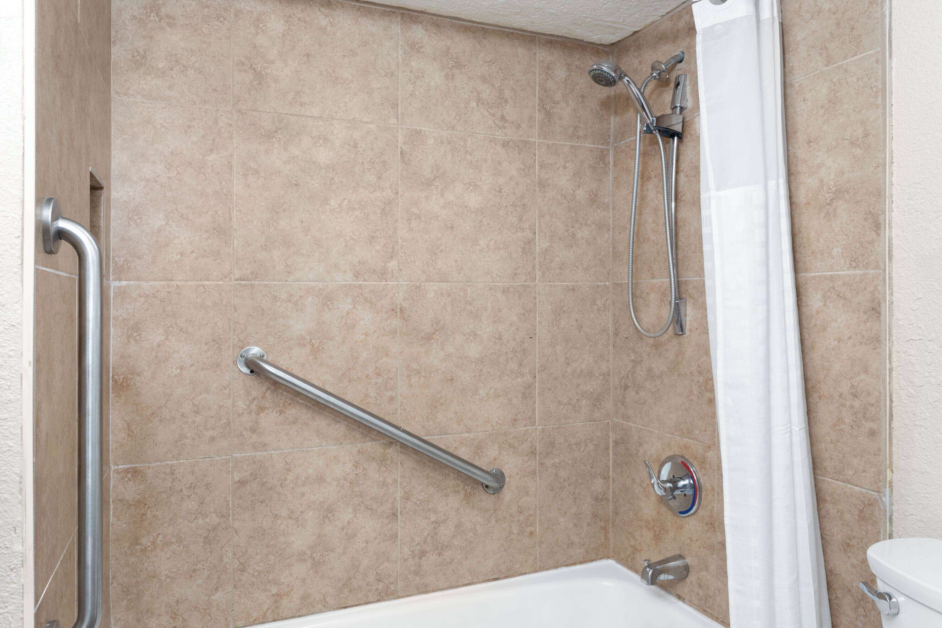 A bathroom with beige tiles, a white bathtub, a shower with a handheld showerhead, and a grab bar.